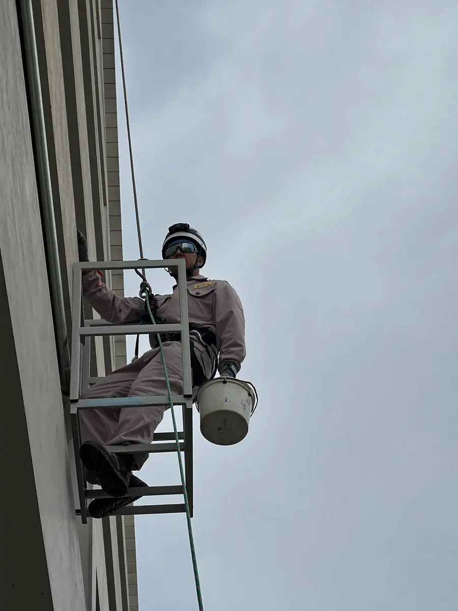 Trabalhador com capacete, cinto e corda de segurança descendo a fachada de um prédio.