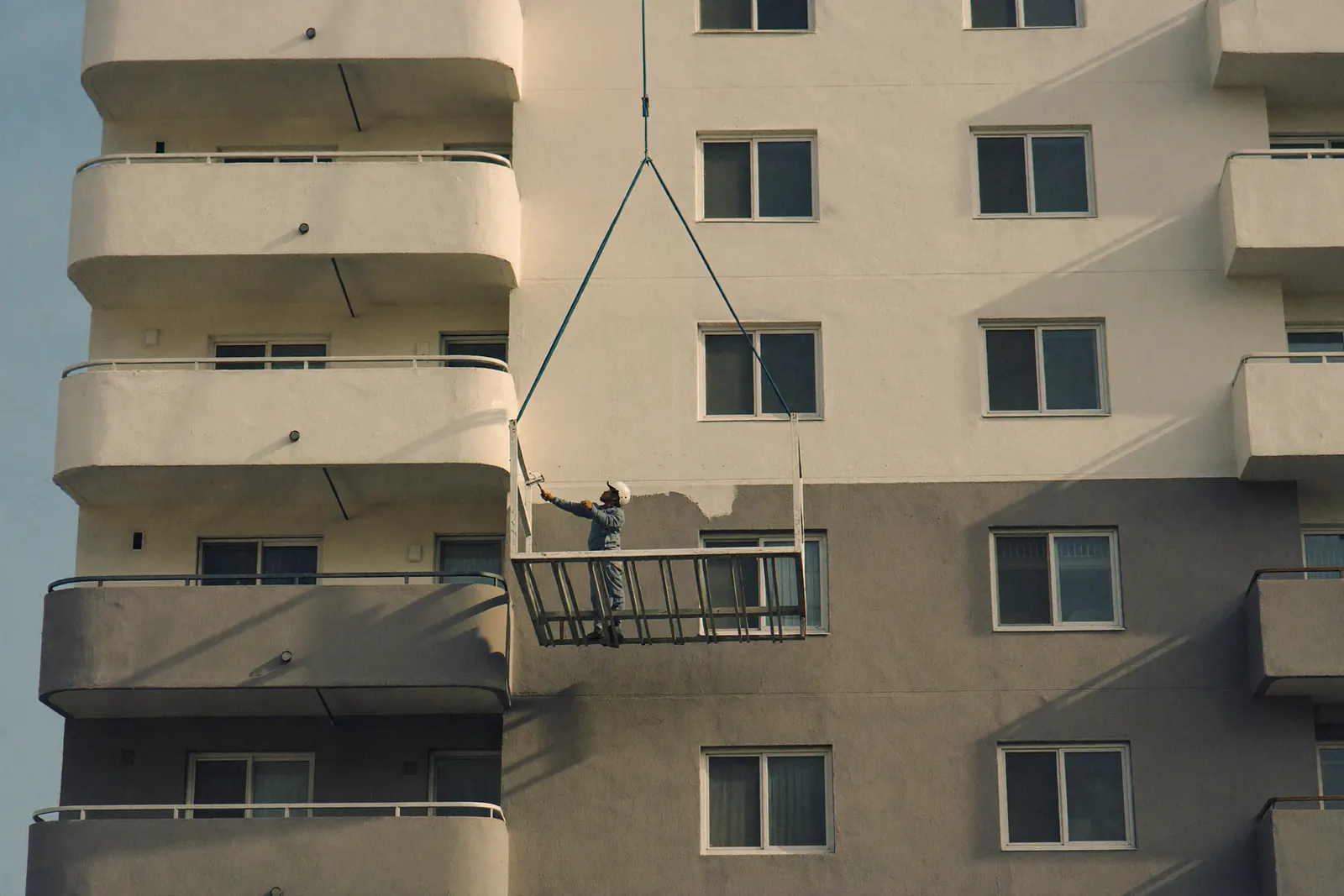 Pintor em balancim pintando a fachada de um prédio residencial; metade de cima já pintada em tom claro.