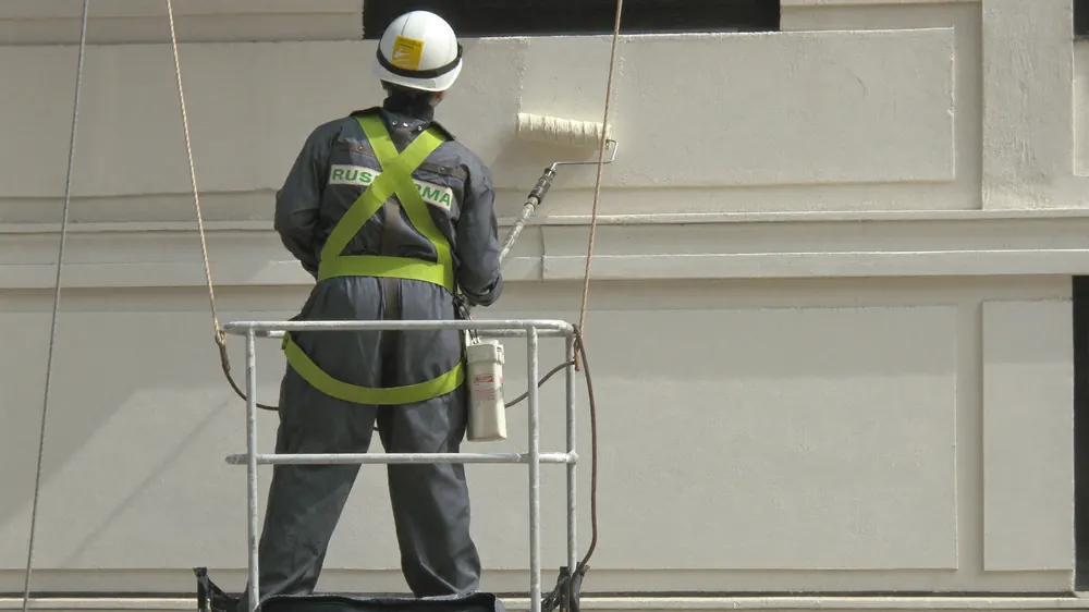 Pintor com cinto de segurança em balancim passando rolo na fachada.