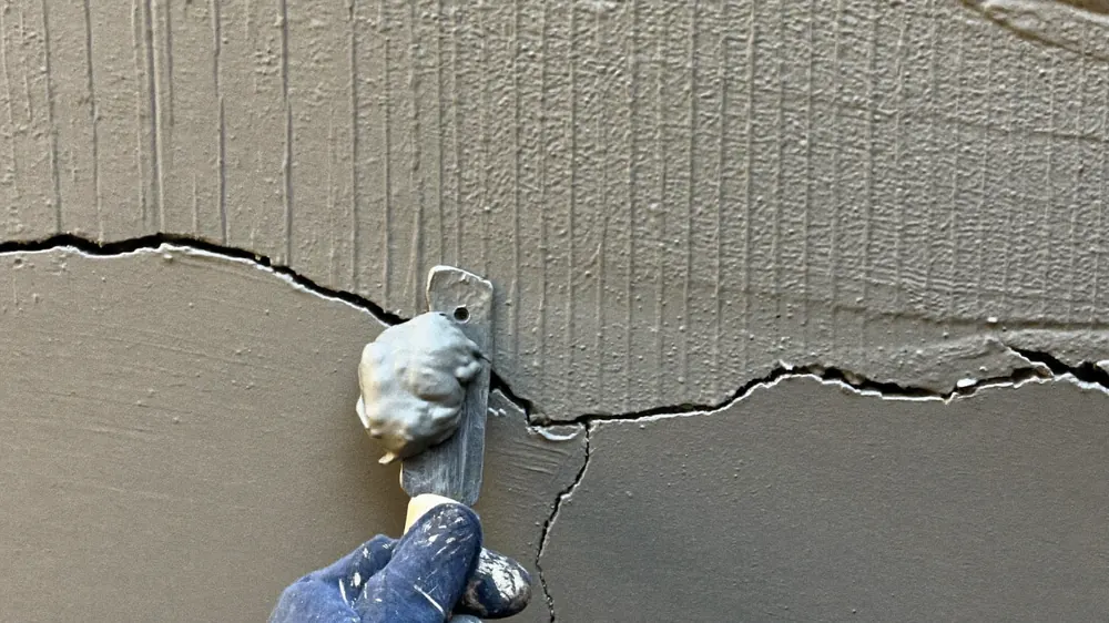 Mão com luva aplicando massa com espátula numa trinca de parede de concreto.